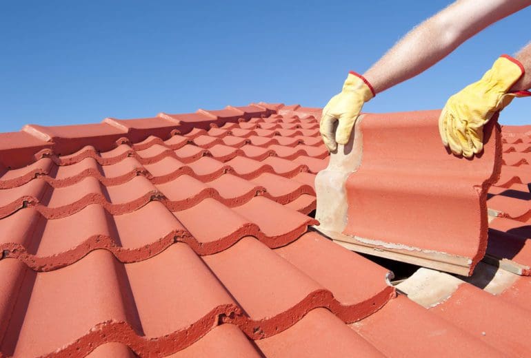 Roof repair worker with yellow gloves replacing red tiles or shingles on house with blue sky as background and copy space.
