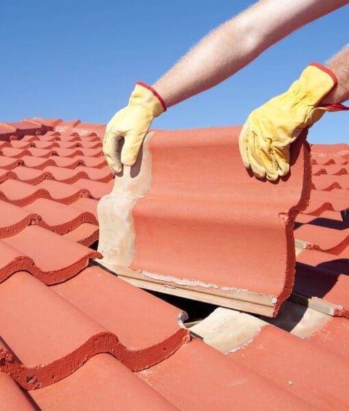 Roof repair worker with yellow gloves replacing red tiles or shingles on house with blue sky as background and copy space.
