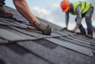 Roofer worker in special protective work wear and gloves, installing asphalt or bitumen shingle on top of the new roof under construction residential building